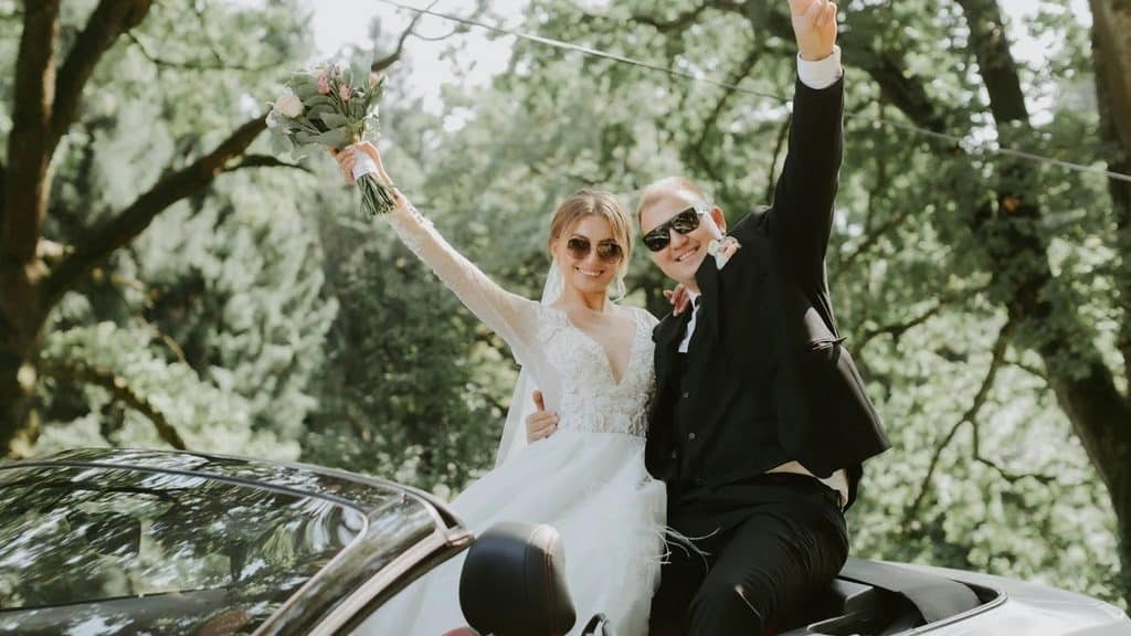 Newlyweds in sunglasses perched on the back of an open convertible, waving as they leave