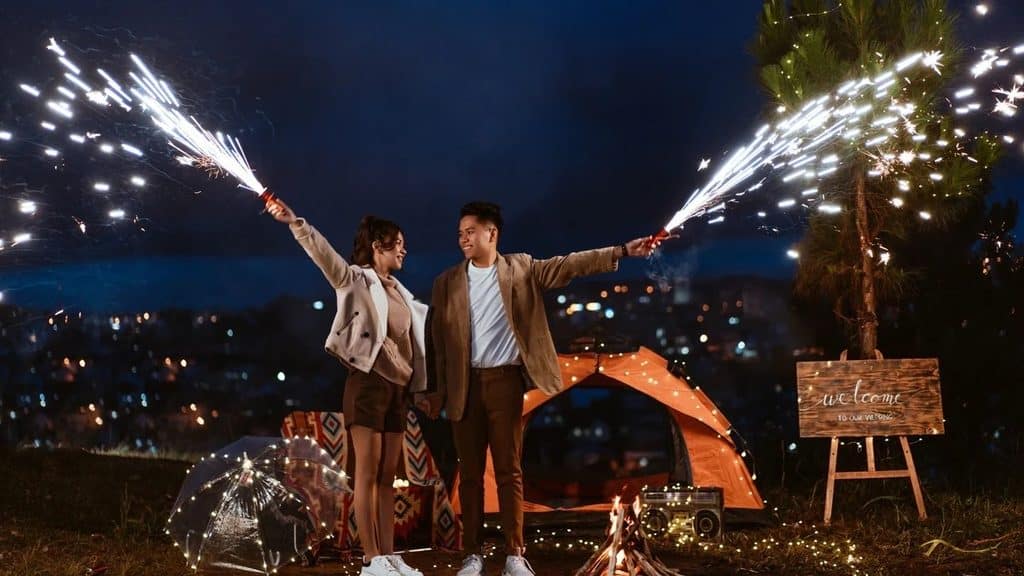 Couple holding sparkler fountains at night beside a wooden welcome sign on an easel